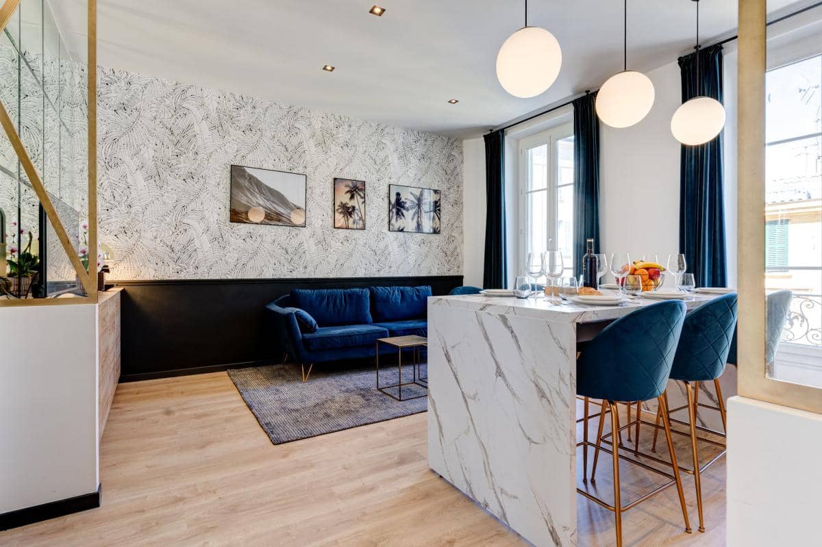 Salon et salle à manger d'un appartement qui proposent des locations. Du marbre blanc, du papier peint tropical noirs et blancs sont très présents. Les meubles sont bleu foncé afin de créer un vrai contraste dans la pièce.